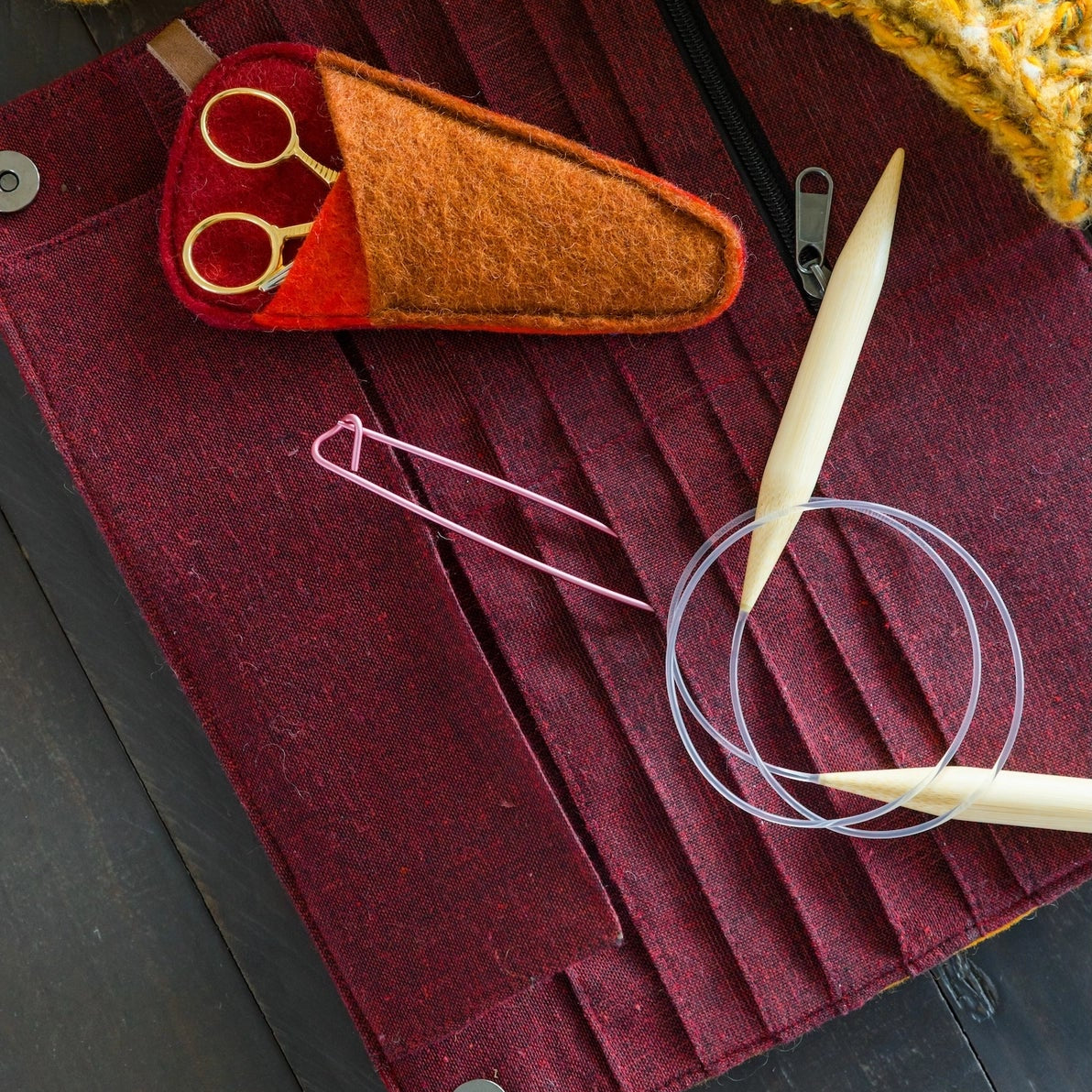 An burgundy needle roll on a dark wooden surface. Visible tools include cream-colored circular knitting needles, a small orange felt pouch with gold-handled scissors, a pink stitch marker, and a metal zipper pull.