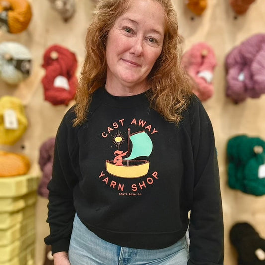 Woman wearing a black sweatshirt with 'Cast Away Yarn Shop' logo in front of yarn balls.