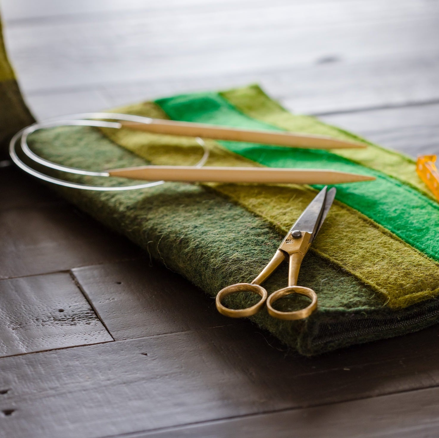 A close-up of a felted wool needle case with green and yellow stripes, containing knitting needles and a gold-handled scissors, resting on a dark wooden table.