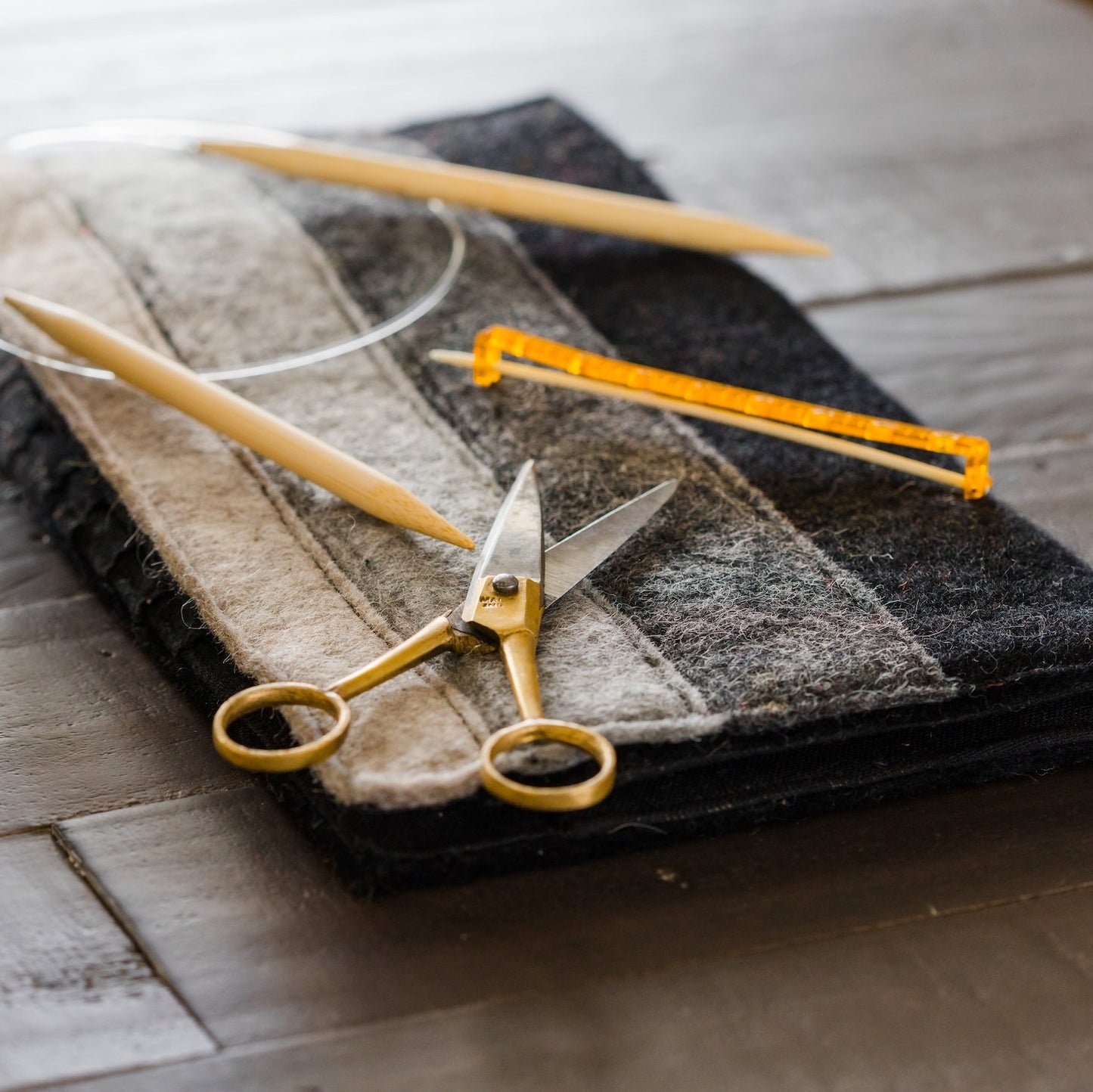 A close-up of knitting or sewing tools arranged on a dark wooden surface: a ball of gray and white yarn, wooden knitting needles, a yellow tape measure, and gold-handled scissors on a folded gray fabric piece.